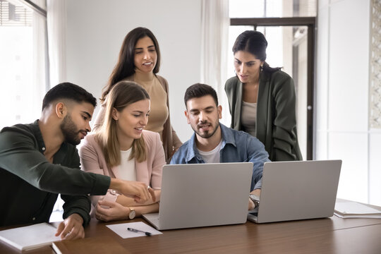 Diverse group of five colleagues take part in collaborative work session gathered around two modern laptops, discussing new AI platform for business, develop start-up strategy. Teamwork, brainstorming
