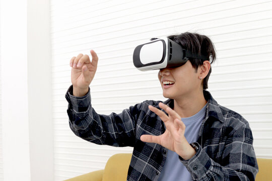 Excited Asian young man with virtual reality glasses raising hands in an empty place to control something on sofa at living room. Teenager man with future modern digital technology at home.