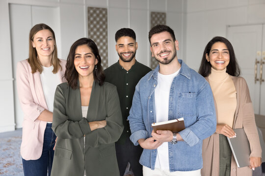 Professional team of five team members posing for camera standing in office, looking at camera with joyful expression, exuding competence, reliability and achievement. Human resources, seminar event