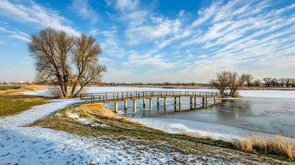 Winter landscape with wooden footbridge over frozen lake, snow-covered banks, trees, and clear blue sky. Ideal for calendars, travel brochures