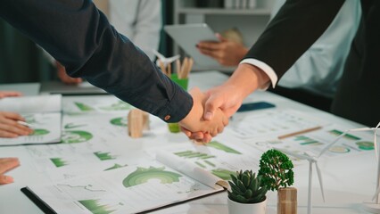 Two business professionals engage in a handshake signaling agreement over financial reports and market analysis in a modern office environment filled with greenery. SACTR