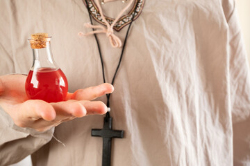 A man with a cross around his neck holding a potion in a red glass bottle