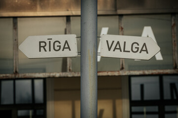 Obraz premium A close-up of a weathered metal signpost pointing to Rīga and Valga, mounted on a pole in front of an old building. The aged backdrop adds a rustic, nostalgic feel.