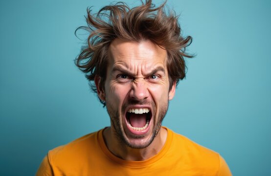 Close-up portrait of man with wild, disheveled hair shows exaggerated angry expression against blue background. Intense emotion, furious, rage, frustration, dramatic aggression. Yellow t-shirt.