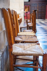 Three beech wood and rush prayer chairs with woven seats are lined up against a stone wall inside the Saint-Paul Saint-Louis Church in Paris, France, creating a sense of order on a summer day.