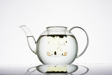 Teekanne aus Glas vor hellem Hintergrund ohne Deckel, gefüllt mit Wasser im Teeeinsatz schwimmt wenig loser Kräutertee, beginnt zu ziehen, Silhouette, minimalistisch