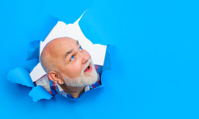 Bearded man with blue color lenses in eyes looking through paper hole. Smiling mature man with...