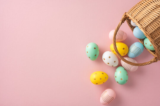 Festive pastel-colored Easter eggs, decorated with various patterns, spill from a wicker basket onto a pink surface, perfect for celebrating spring or Easter-themed concepts