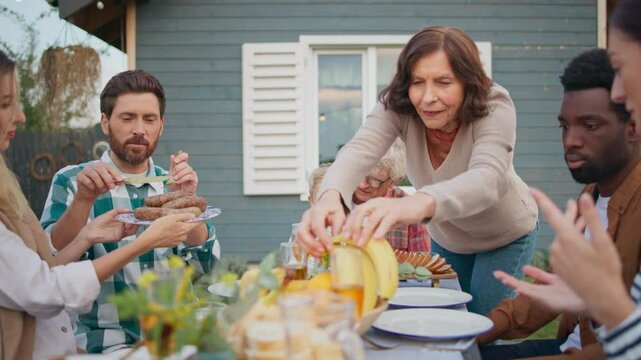 Friendly family sitting at table and enjoying delicious food. Helping each other by passing dishes from other side of table. Smiling and talking while having dinner together in fresh air.