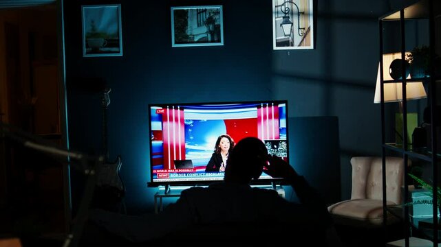 Relaxed person watching breaking news on television at night, chilling on sofa in living room and watching tv program. Young adult listening to media information on global network. Camera A.