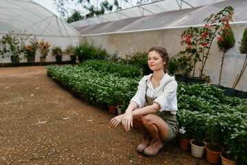 Young woman in garden kneeling among green plants, wearing casual attire and smiling, showcasing...