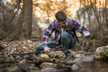 man hiker squats by a creek filling a mug with water from creek