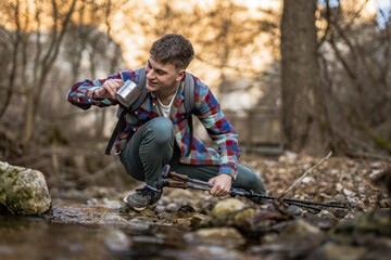 man hiker squats by a creek filling a mug with water from creek