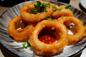 A plate holds several golden-brown, crispy onion rings garnished with fresh cilantro and green onions, accompanied by a spicy dipping sauce