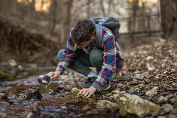 man hiker squats by a creek filling a mug with water from creek