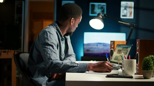 African American freelancer working from home writing notes and researching data driven topics. Focused on productivity and planning tasks in a modern apartment for remote work success. Camera A.