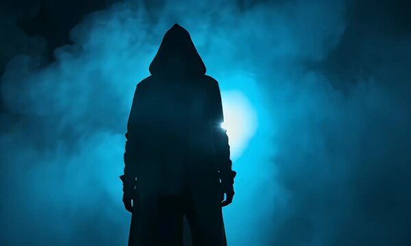 Silhouette of Hooded Figure in Dark Cloak Standing in Blue Smoky Backlit Background Creating Mysterious and Ominous Atmosphere