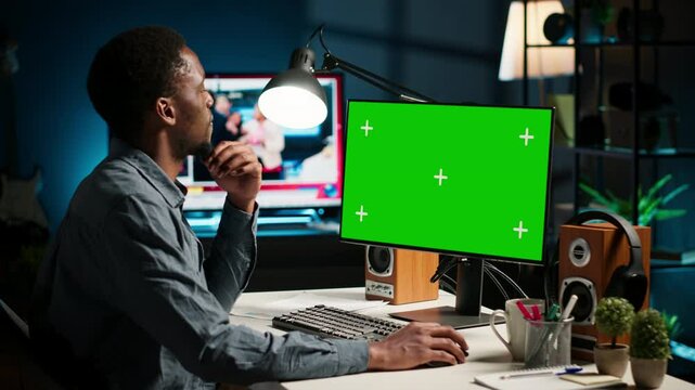 African american person working from home and checking green screen on PC, enjoys remote job at a home office setup. Teleworker multitasking and networking in his modern loft. Camera B.