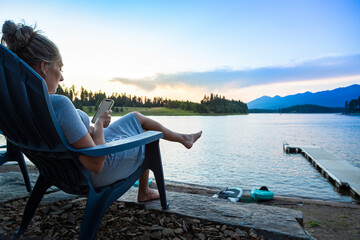 Woman relaxing from her lakeside home overlooking a scenic lake in the Montana mountains. She is sitting and looking at her smartphone with a beautiful sunset in the background.