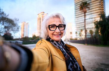 Elegant senior woman taking selfie looking at camera with stylish glasses and a yellow jacket walks...