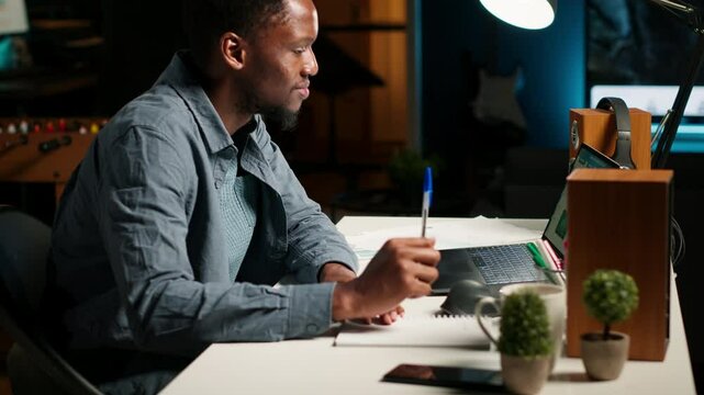 Self employed Black man multitasking at home taking handwritten notes and browsing the web for project research. Managing schedule and tasks with a planner to boost productivity. Camera B.