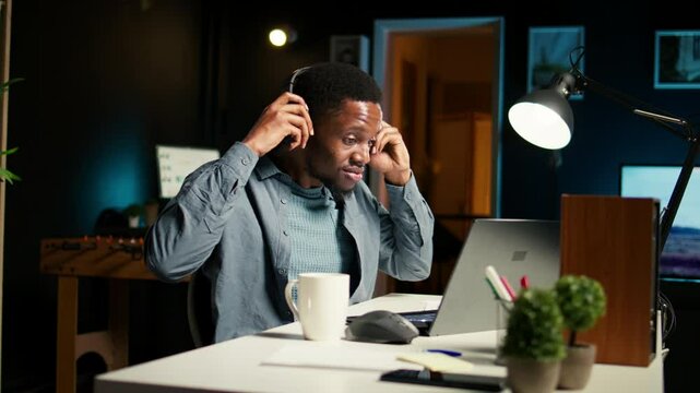 African american specialist closing the monthly balance with a show on headset, working remotely while he listens to his favorite podcast. Managing expenses and insurances from home. Camera A.