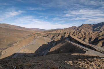 A winding road cuts through the rugged landscape of the Atlas Mountains in Morocco, under a partly cloudy sky.