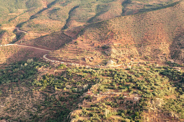 A panoramic view of the majestic Atlas Mountains in Morocco, contrasting against a clear blue sky. Lush green valleys wind through the rugged terrain,.