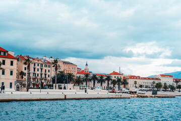 Obraz premium Scenic waterfront view of a historic European coastal city with palm trees, colorful buildings and a cloudy sky. Split, Croatia