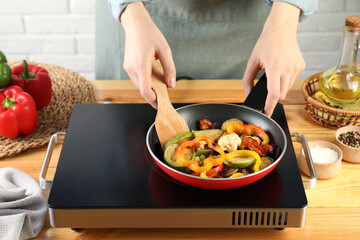 Woman frying vegetables in pan on stove at wooden table indoors, closeup