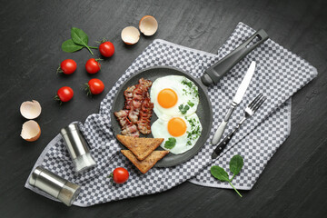 Tasty fried eggs, bacon and pieces of toast served on black table, flat lay