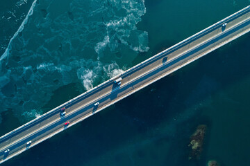 Aerial top down view of a cars driving along a coastal bridge with clear waters and ocean waves beneath in bright daylight