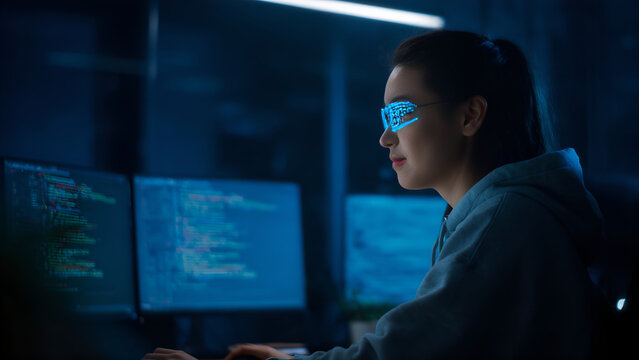 Young Programmer in Hoodie Coding Software with AR Glasses Displaying Data in a Dark Office.