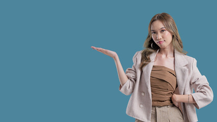 A woman is standing in front of a blue background, wearing a tan jacket and tan pants. She is...