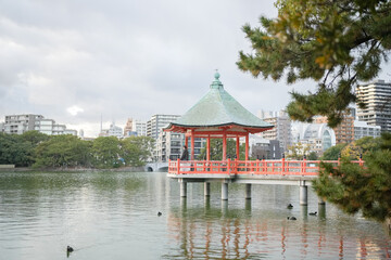 Winter at Ohori Park in Chuo Ward, Fukuoka City, Japan. A 29-year-old Japanese woman wearing a checkered coat. She watches the sunset on a traditional red structure floating in a pond.