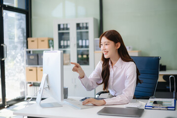 Obraz premium Asian woman, smiling and working at her desk in an office environment. She is wearing a business suit and holding documents