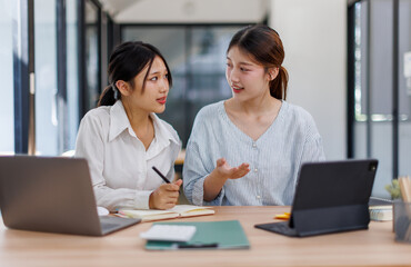 Obraz premium Happy professional young women collaborating with enthusiasm in a modern office setting. Two asian female entrepreneurs using a laptop for a productive business meeting in a corporate workplace. 