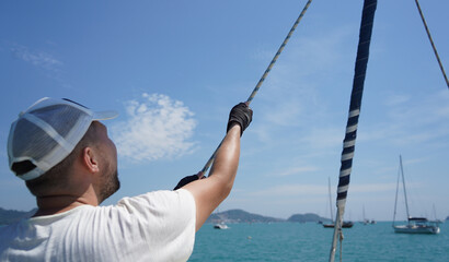 Male captain on deck of sailboat opening sails pulling the rope