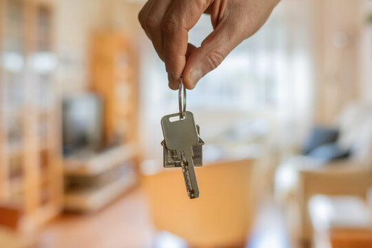 Hand holding house keys with house shaped keychain in front of blurred new home background, symbolizing real estate investment, mortgage, and property ownership