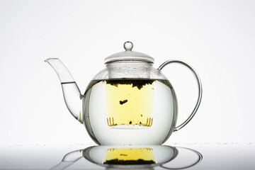Teekanne aus Glas vor hellem Hintergrund mit Deckel, gefüllt mit Wasser, im Teeeinsatz schwimmt loser Kräutertee und färbt das Wasser gelb, wenig gezogen, Silhouette, minimalistisch