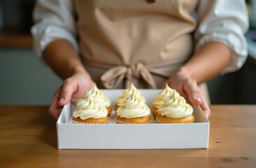 Close up view of woman hands holding delivery box with cupcakes decorated with  light yellow whipped cream. Pastry chef is wearing light beige apron. Confectionery and pastry business, self occupation