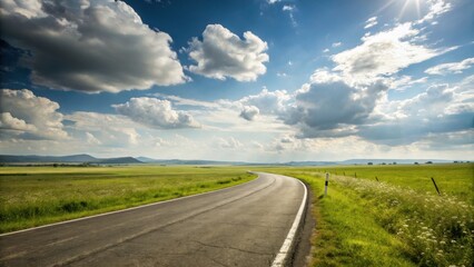 Fototapeta premium A winding asphalt road leads through a vast green field under a bright blue sky with fluffy clouds, sunlight filtering through the clouds, casting a warm glow on the scene.