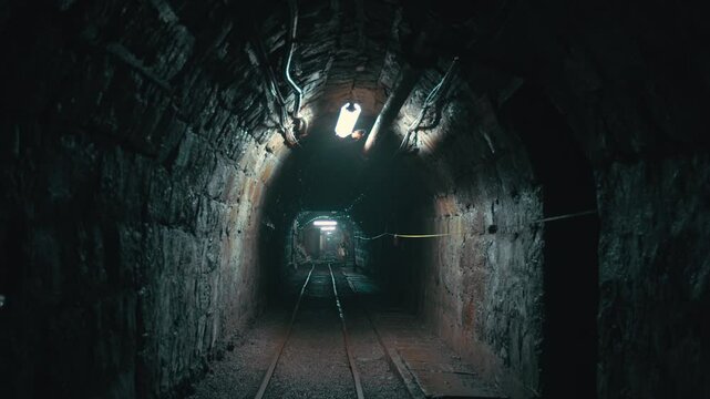 An old mine tunnel. The weak lights on the ceiling reflect on the damp stone walls and create a dim atmosphere. The cold and abandoned feeling of the environment is reflected to the viewer.
