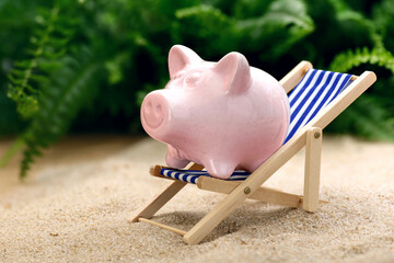 Piggy bank in a beach chair on sand with tropical green plants in the background, concept for saving money when booking a holiday, on vacation or on a trip