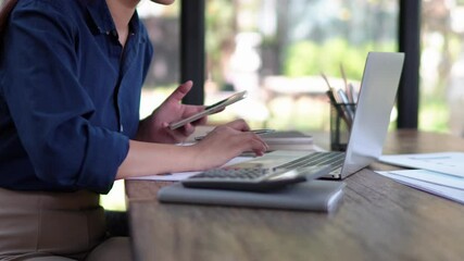 Focused businesswoman using smartphone and laptop for financial analysis in a natural-themed office environment.