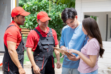 Young couple pointing and discussing moving logistics with movers in red uniforms, standing near truck filled with moving boxes. Movers appear focused, with a clipboard showing transportation fee