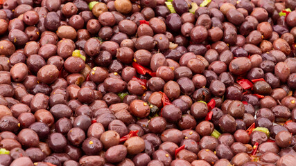 Background of pickled dark olives combined with red chili pieces and green herbs reflecting a shiny oily surface and soft texture
