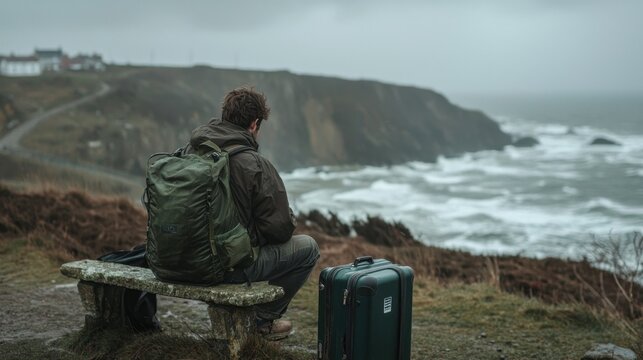 A traveler sits alone on a stone bench overlooking rough ocean waves. The cloudy sky adds to the solitary atmosphere, as luggage rests nearby, suggesting a journey