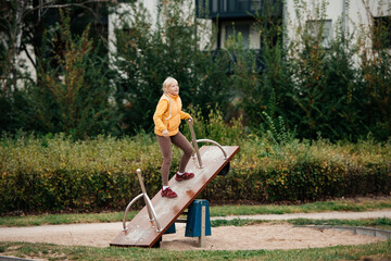 A blonde-haired girl in bright clothes balancing on a seesaw, embracing carefree childhood.