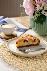 A slice of almond and berry cake on a white plate, accompanied by a cup of cappuccino, flowers, and a cozy table setting, creating a warm and inviting atmosphere.